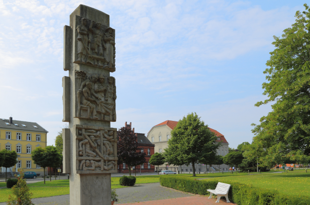 Ein steinernes Denkmal steht in einem Park mit einer Bank davor, umgeben von grünem Gras, Pflanzen und Bäumen, mit Gebäuden, Fahrzeugen und einem bewölkten Himmel im Hintergrund.