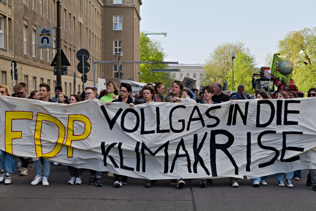 Eine Gruppe von Menschen marschiert mit einem Banner "FDP Vollgas in die Klimakrise" die Straße entlang, mit Gebäuden, Bäumen und Verkehrszeichen im Hintergrund unter einem klaren blauen Himmel.