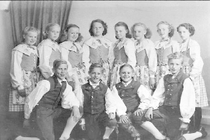 Schwarzes und weißes Foto von Kindern, die vor einer Vorhangwand posieren, mit dem Text "1930er Kinder-Schule" unten.