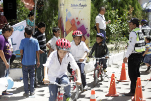 Gruppe von Kindern auf Fahrrädern auf einer Straße mit Verkehrshütchen, einige tragen Helme, andere stehen in der Nähe, mit einem Banner, Bäumen und Gebäuden im Hintergrund.