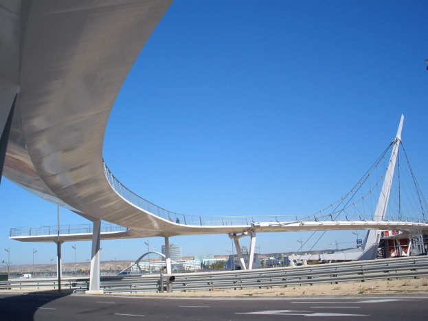 Große Brücke mit Geländern, Pfählen und Laternen, die eine befahrene Autobahn überspannt, mit Gebäuden, Bäumen und einem klaren blauen Himmel im Hintergrund.