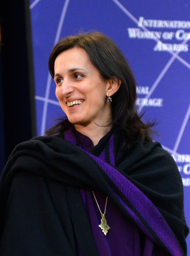 Eine Frau in einem lila Kleid und schwarzer Schärpe steht neben einer anderen lachenden Frau, mit einem Banner im Hintergrund, auf dem 'Internationale Frauen der Farbe Awards' steht.