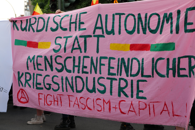 Eine Gruppe von Menschen steht auf der Straße und hält ein pinkes Banner mit der Aufschrift "Kurdische Autonomie Statt Menschenfeindlicher Kriegsindustrie Fight Fascism Capital", mit Ampeln, Flaggen, Bäumen, Gebäuden und einem klaren blauen Himmel im Hintergrund.