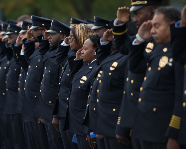 Eine Gruppe von Polizisten in Uniform steht in einer Reihe und salutiert vor einem unscharfen Hintergrund von Bäumen.