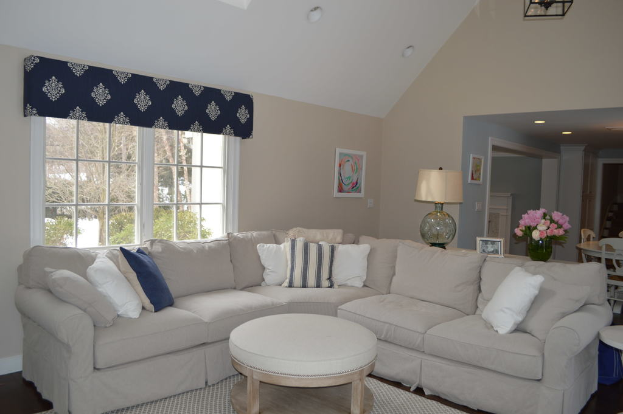 Ein Wohnzimmer mit einer Couch, Kissen, einem Tisch, Stühlen, einem Blumenstrauß, einem Foto Rahmen, einem Wecker und einem Fenster auf der linken Seite mit an der Decke angebrachten Lampen.