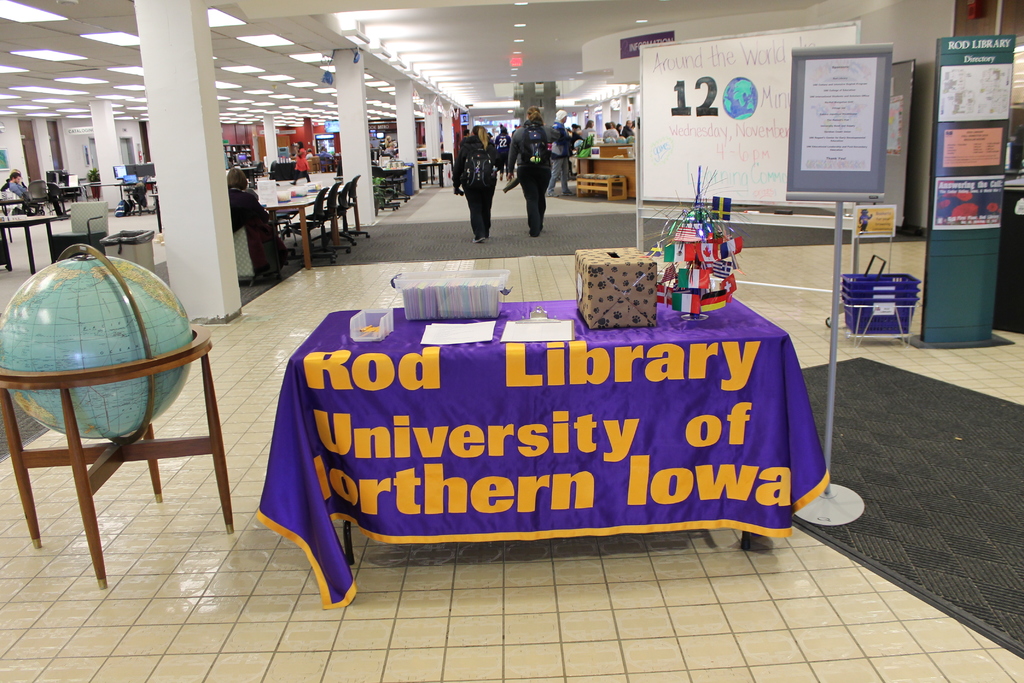 Eine Ausstellung mit einem Tisch, auf dem verschiedene Landesflaggen, eine Schachtel, Papiere und ein Banner sowie eine Kugel links und ein Anschlagbrett rechts mit einigen Menschen in der Nähe und mehreren Tischen und Stühlen im Hintergrund zu sehen sind.