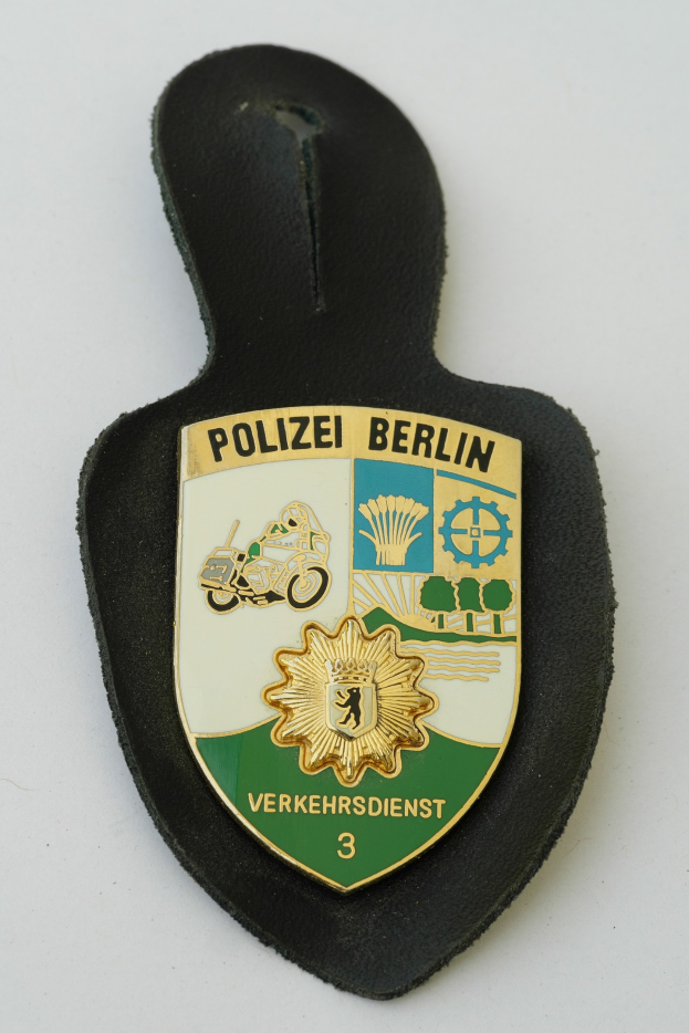 Ein schildförmiges Polizeibadge mit 'Polizei Berlin' in fetter schwarzer Schrift auf einem blauen Hintergrund mit einem weißen Stern und Rahmen, das auf einer Oberfläche ruht.