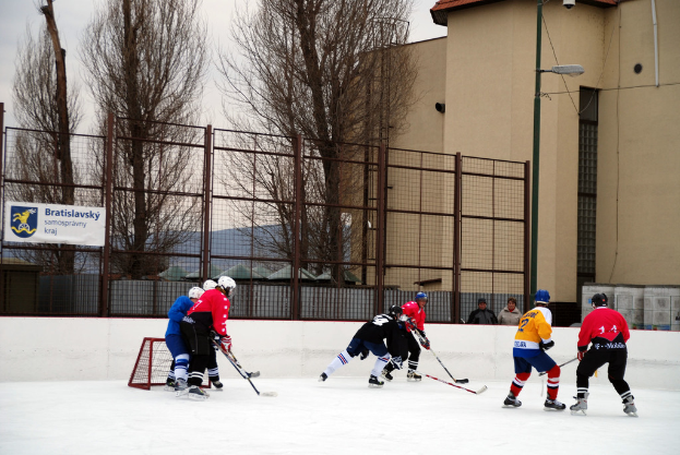 Menschen beim Eishockeyspielen auf einem Eisplatz mit Gebäuden, Bäumen, einer Straßenlaterne, einem Namensschild und Zäunen im Hintergrund unter einem Himmel.