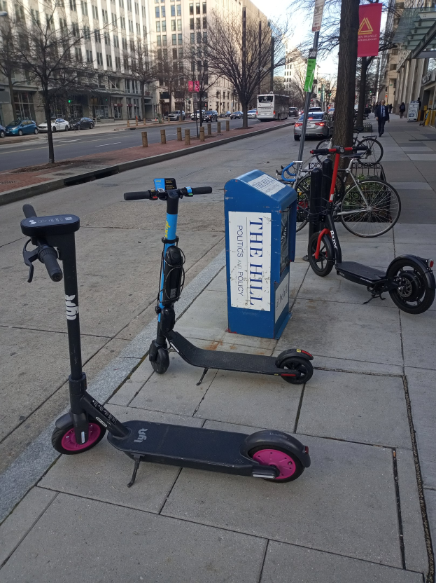 Zwei Elektro-Scooter auf einer Stadtstraße mit Fahrrädern, einem Müllcontainer, Fahrzeugen, Bäumen, Gebäuden und Menschen im Hintergrund.