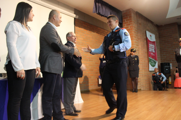 Eine Gruppe von Menschen auf einer Bühne, einer in Uniform mit einer Mütze, eine Frau in einem weißen Hemd, mit einem Banner, Lautsprecher, Vorhöngen, Tisch und Wandbildschirm im Hintergrund.