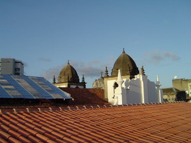 Stadtansicht mit Gebäuden im Vordergrund, einem blauen Himmel im Hintergrund und Solarpanels auf einem Dach, die den Einsatz erneuerbarer Energien anzeigen.