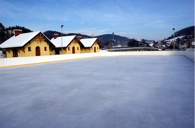Eine Eislaufbahn in einem verschneiten Feld umgeben von Häusern, Laternen, einem Zaun, Bäumen, Hügeln und einem bewölkten Himmel.