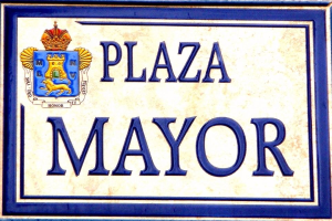 Rechteckige weiße Plakette mit schwarzem Text "Plaza Mayor" und einem blauen Rahmen, die ein gekröntes Wappen-Logo zeigt, an einem Gebäude angebracht.