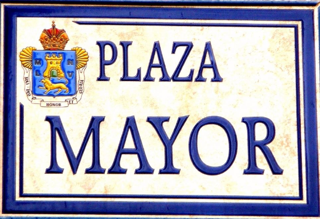 Rechteckige weiße Plakette mit schwarzem Text "Plaza Mayor" und einem blauen Rahmen, die ein gekröntes Wappen-Logo zeigt, an einem Gebäude angebracht.