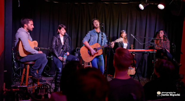 Fünf Personen auf der Bühne, zwei spielen Gitarren, alle mit Mikrofonen auf Ständern, mit einem Vorhang und Scheinwerfern im Hintergrund.