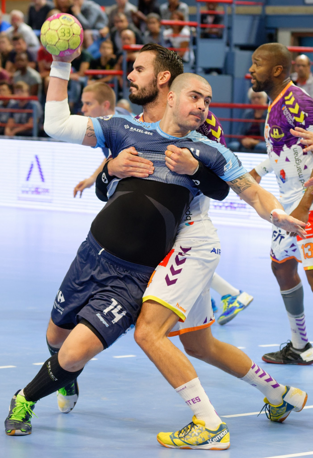 Männer spielen Hallenhandball, einer hält den Ball, mit Zuschauern auf Stühlen, Geländern und einer Wand in einer Stadionatmosphäre.