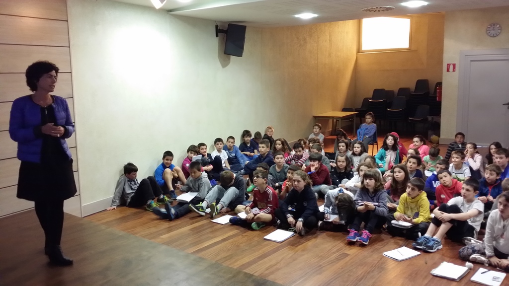 Kinder auf dem Boden sitzend mit Büchern vor sich, eine Frau auf der Bühne links stehend und eine Wand im Hintergrund.