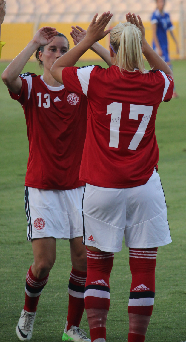 Zwei Frauen in roten T-Shirts und weißen Shorts stehen auf einem Fussballfeld und klatschen in die Hände, mit einer Person in einem blauen Kleid, einer gelben Tafel und ein paar Stühlen im Hintergrund.