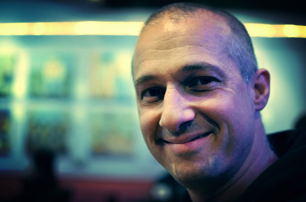 Ein Mann in einem schwarzen Kleid lacht vor einem unscharfen, hell erleuchteten Hintergrund.