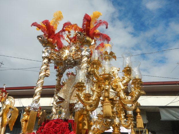 Ein großes goldenes und rotes Festwagen, das mit Blumen und Dekorationen geschmückt ist, bei einem Karnevalsumzug, mit einem Gebäude, Strommasten mit Drähten und einem bewölkten Himmel im Hintergrund.