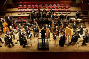 Großes Orchester auf der Bühne mit Dirigent, Musikern mit verschiedenen Instrumenten, Stühlen und Hintergrundtreppe.
