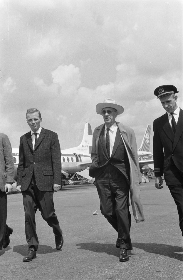 Eine Gruppe von Männern in Anzügen und Hüten geht auf ein Flugzeug auf dem Runway zu, mit einem klaren Himmel im Hintergrund, dargestellt in Schwarz-Weiß.