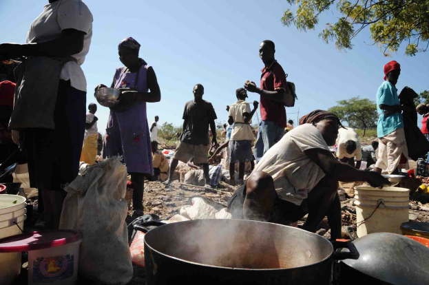 Eine Gruppe von Menschen, die in einer ländlichen Gegend Essen zubereiten, mit einem großen Topf, Eimern und Abdeckungen auf dem Boden, unter einem klaren blauen Himmel.