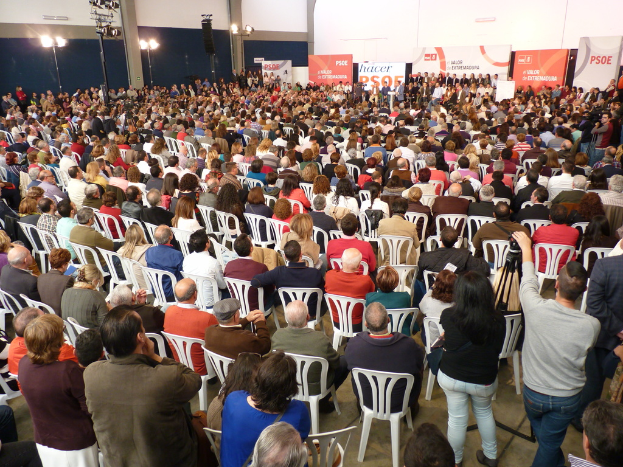 Ein volles Stadion mit vielen sitzenden und stehenden Menschen, eine Bühne mit zwei Rednern am Podium und verschiedene Banner und Lichter.