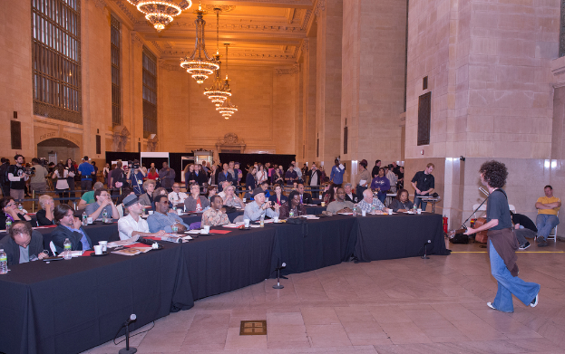 Eine Konzertszenen mit einer Menge Menschen, einem Mann, der auf der rechten Seite Gitarre spielt und singt, einer Wand und Fenstern im Hintergrund, Lichtern oben und einem Boden unten.