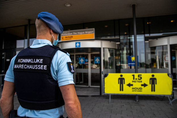 Ein Polizist in einer blauen Uniform und Mütze steht vor einem gläsernen Gebäude mit einer gelben Tafel auf der rechten Seite und zusätzlichen Tafeln im Hintergrund.