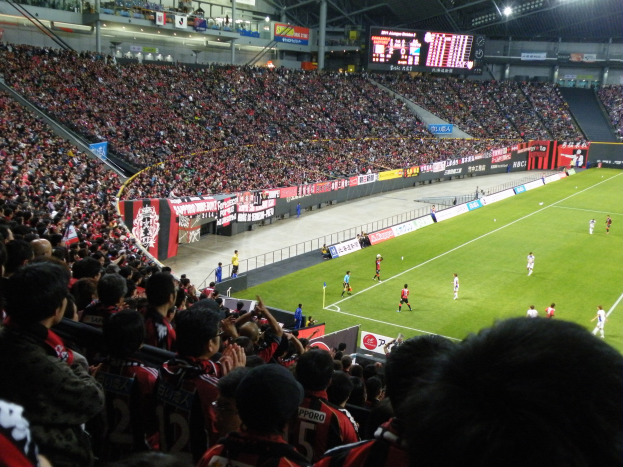 Ein Fußballspiel in einem großen Stadion mit Spielern auf dem Feld und Zuschauern in den Rängen, mit Bannern, einem Zaun, einem Anzeigebildschirm, Metallstangen und Deckenleuchten.