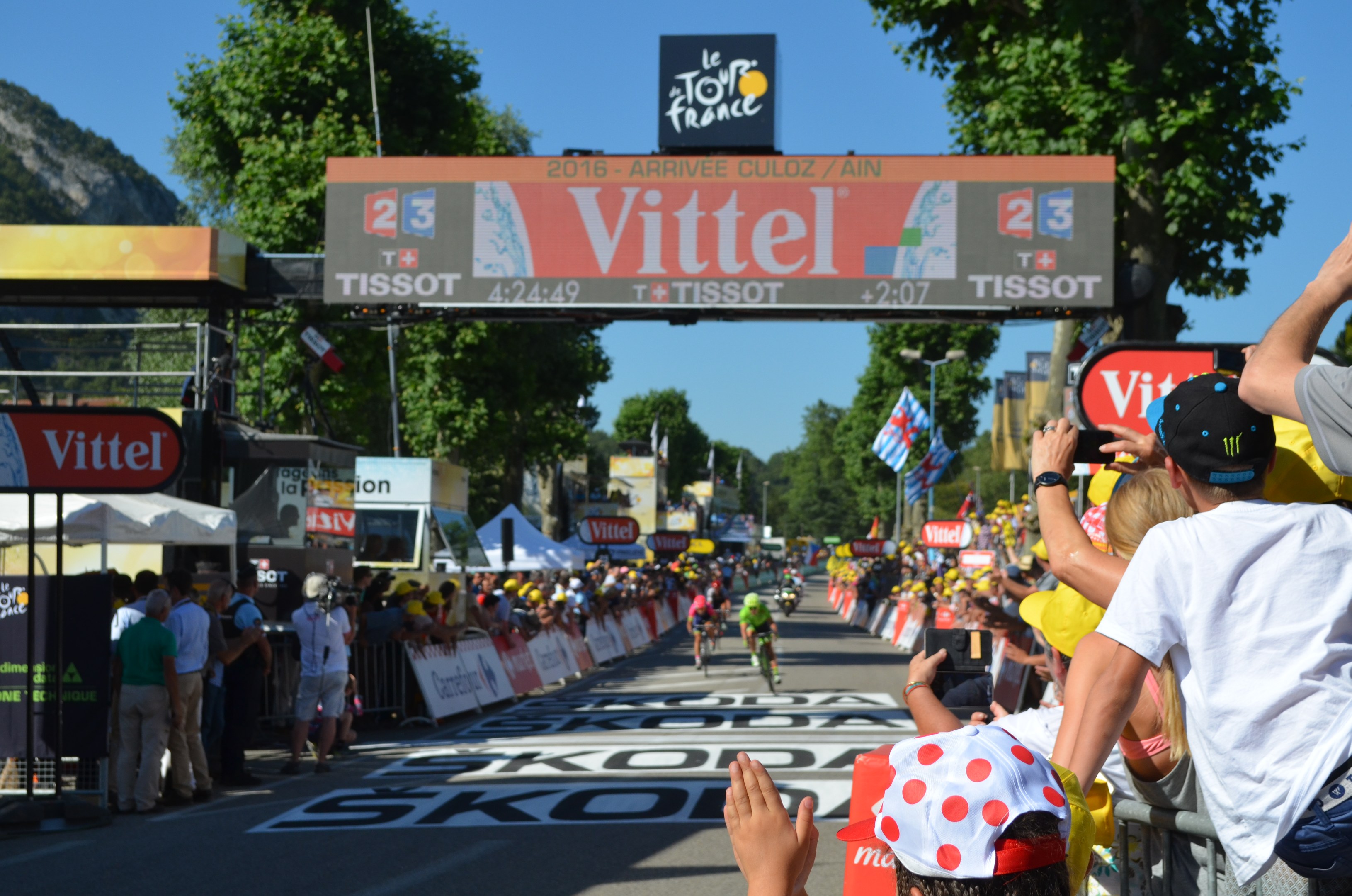 Fahrradfahrer fahren unter einem Bogen mit der Aufschrift "Tour de France 2016" eine Straße entlang, mit Zuschauern an den Seiten und Bäumen, einem Hügel und einem klaren blauen Himmel im Hintergrund.