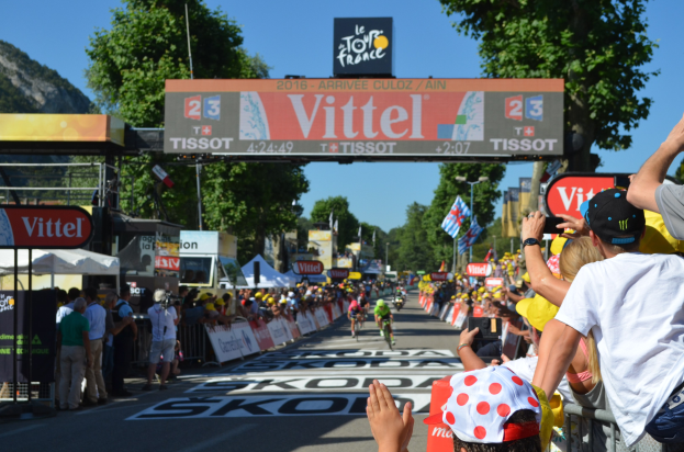 Fahrradfahrer fahren unter einem Bogen mit der Aufschrift "Tour de France 2016" eine Straße entlang, mit Zuschauern an den Seiten und Bäumen, einem Hügel und einem klaren blauen Himmel im Hintergrund.