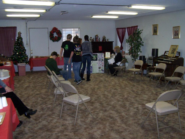 Ein Raum mit mehreren Stühlen, Tischen und Menschen, einige stehen, andere sitzen, mit Tassen und Gefäßen auf den Tischen, einem Weihnachtsbaum, einem Piano auf der rechten Seite, Foto Rahmen, Fenstern und Röhrenleuchten an der Decke sowie einer Wand mit einem Strauß, Vorhang, Tür und weiteren Foto Rahmen im Hintergrund.