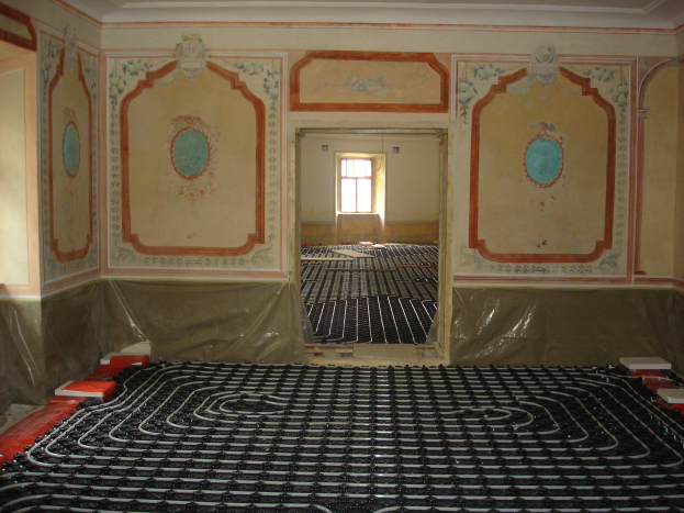 Ein Raum mit einer Fußbodenheizung in der Mitte, umgeben von Wänden und einem Fenster im Hintergrund, mit verschiedenen Gegenständen auf dem Boden, die wahrscheinlich mit dem Heizsystem zusammenhängen.