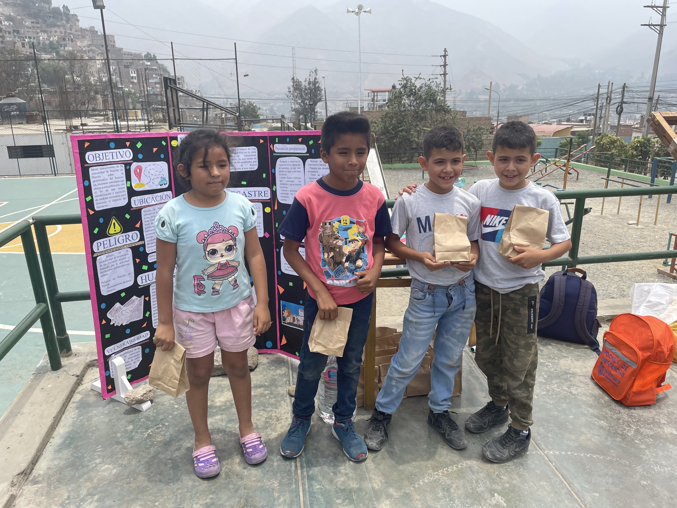 Eine Gruppe von Kindern steht auf einem Basketballfeld und hält Papiertüten in den Händen, mit Tüten und Gegenständen rechts und einer Tafel mit Text links, vor einem Hintergrund aus Zaun, Pfosten, Straßenlaternen, Bäumen, Gebäuden, Hügeln und Himmel.