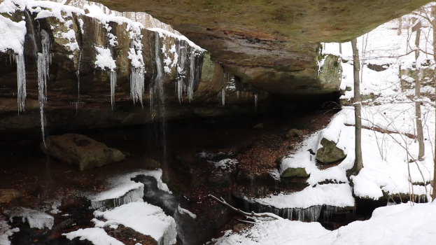Ein kleiner Wasserfall ergießt sich eine schneebedeckte, felsige Klippe in einem bewaldeten Gebiet hinab, mit Eiszapfen an den Felsen und schneebedeckten Bäumen im Hintergrund.