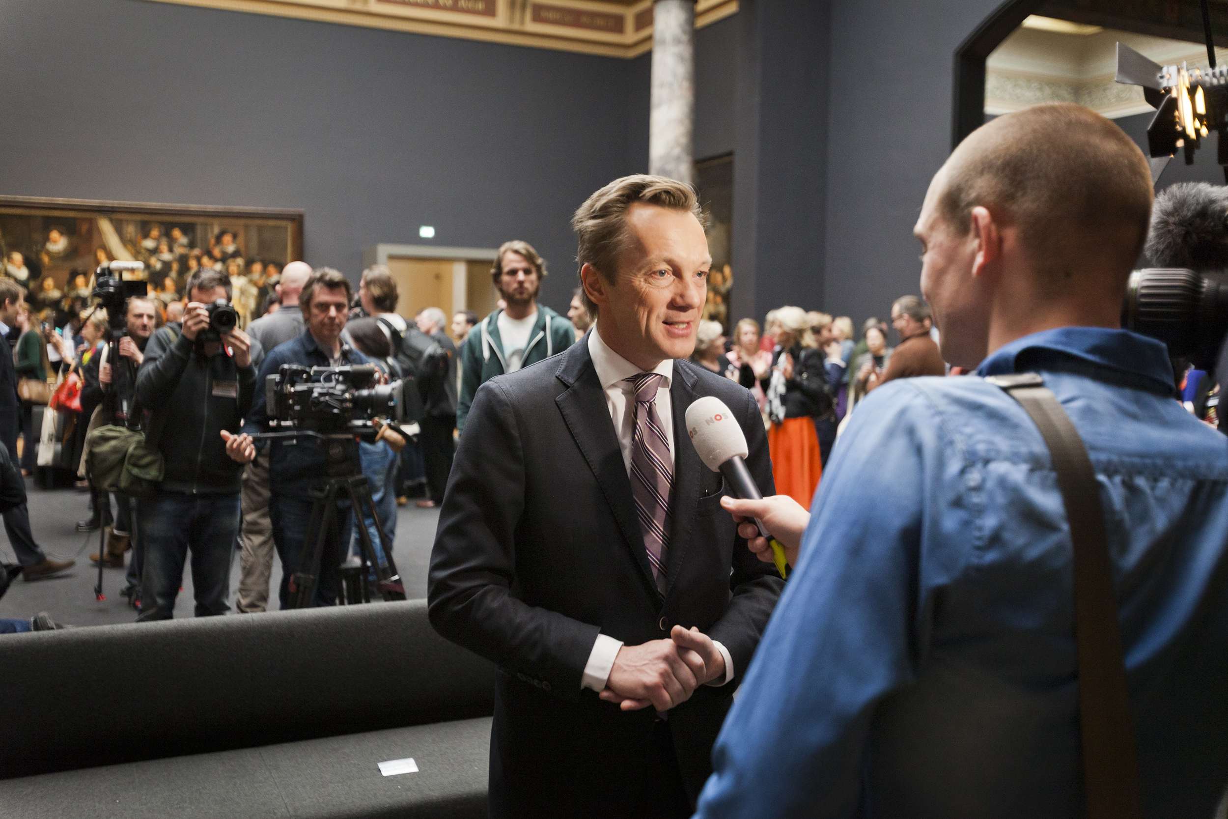 Ein Mann im Anzug und Krawatte spricht mit einem Reporter vor einer Menge, einige halten Kameras, mit einem Sofa im Vordergrund und einer Wand mit einem Foto, Lampen und einer Säule im Hintergrund, hält ein Mikrofon.