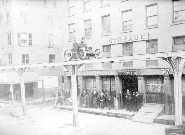 Altes Schwarz-Weiß-Foto eines Feuerwehrautos vor einem Gebäude mit einer Namensplakette, einer Person auf dem Auto, einer Gruppe von Menschen in der Nähe und einem Hund auf dem Gehweg.