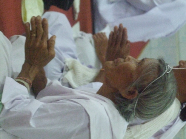 Eine ältere Frau liegt in einem Krankenhausbett mit gefalteten Händen, die besorgt wirkt, mit geschlossenen Augen und Kopf auf den Händen ruhend, umgeben von ein paar Menschen im Hintergrund.