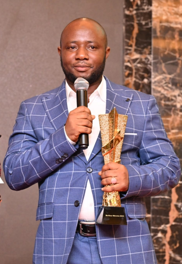 Ein Mann in einem blauen Anzug hält ein Mikrofon und eine Trophäe und steht selbstbewusst mit einem freudigen Gesichtsausdruck da, während er seinen Erfolg gegen eine Wand im Hintergrund feiert.