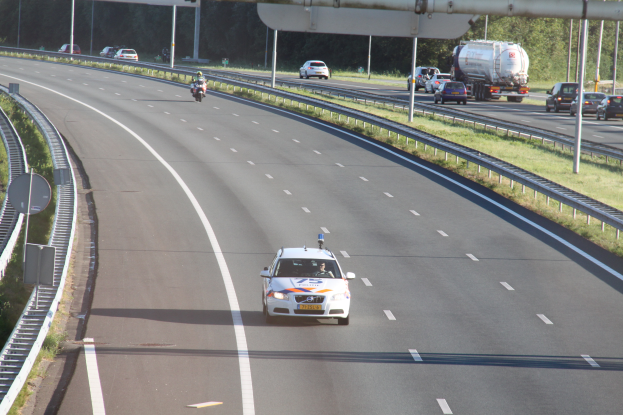 Ein Auto fährt auf einer Autobahn mit einem Polizeiwagen am Rande, anderen Fahrzeugen, Gelöndern, Pfosten, Planken und Bäumen im Hintergrund.