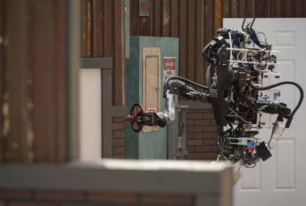 Ein humanoider Roboter mit ausgestreckten Armen und leicht geneigtem Kopf steht vor einem Gebäude mit einer Tür und einer Wand im Hintergrund.