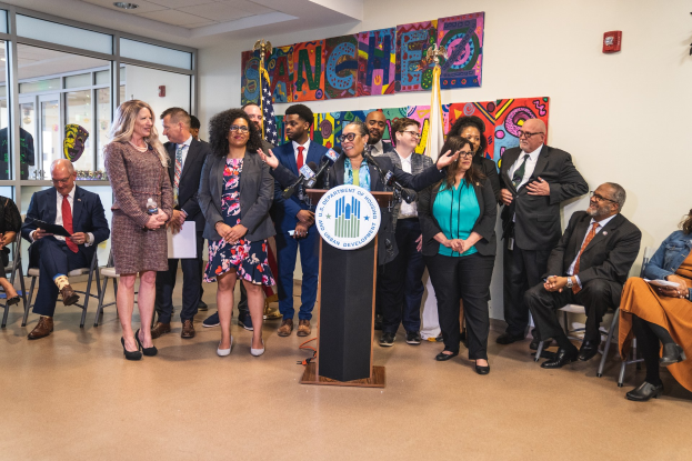 Gruppe von Menschen vor einem Rednerpult mit Mikrofon und einer Tafel mit Text, einige sitzen auf Stühlen und andere stehen, mit Flaggen, einer Tafel, einem Feuerlöscher und Deckenleuchten an der Wand.