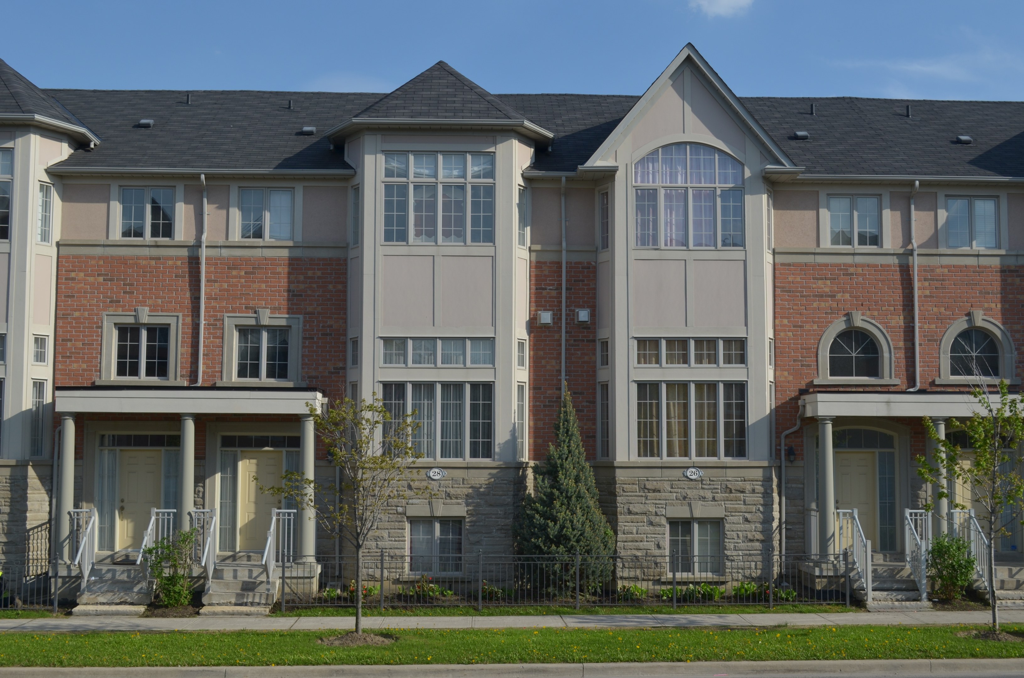 Eine Reihe von Stadthäusern in einem Wohnviertel mit sichtbaren Fenstern, Türen, Säulen, Geländern, Stufen, Pflanzen, Bäumen, Gras, einer Straße im Vordergrund und dem Himmel im Hintergrund.