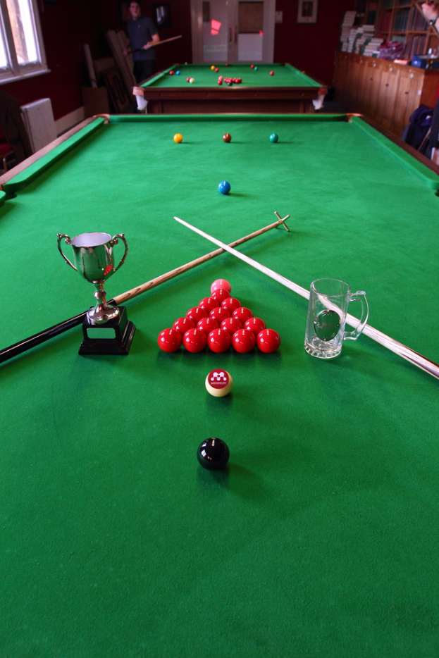 Ein Pokal, ein Glasbecher und mehrere Bälle sind auf einem Billardtisch arrangiert.