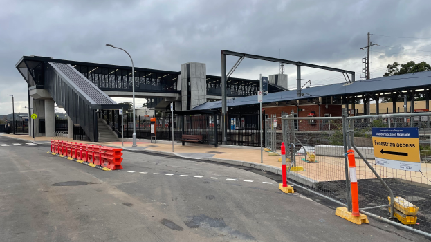 Ein Bahnhofsgebäude mit einer Brücke im Hintergrund, Straßenmasten, Straßenlaternen, Strommasten, Stromkabel, Bäume, Gebäude, Fahrzeuge auf der Straße, Absperrmasten, Schilder, Bänke und ein bewölkter Himmel.
