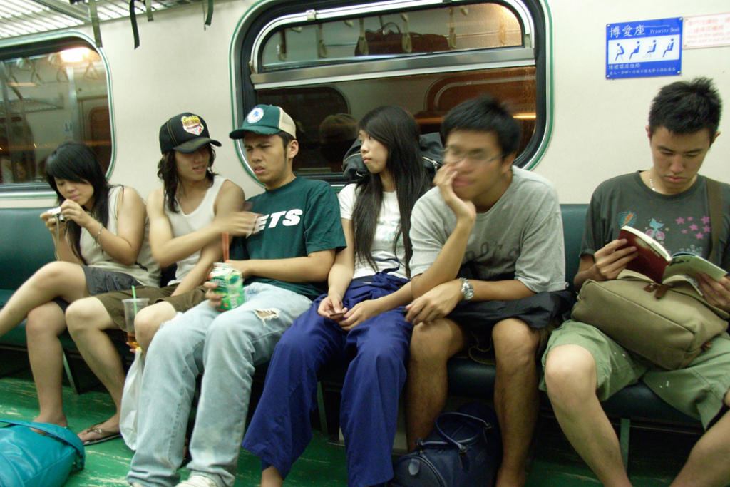 Menschen sitzen auf Sitzen in einem U-Bahn-Zug, mit Glasfenstern im Hintergrund; eine Person hält ein Buch, eine andere eine Tetra-Packung, eine andere eine Dose und eine andere eine Kamera, während Taschen auf dem Boden plaziert sind.