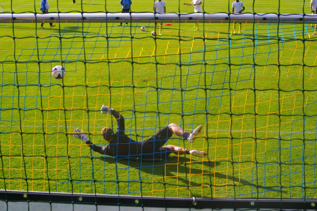 Ein Fussballspieler taucht ab, um einen Ball vor dem Tor zu halten, mit einem Netz im Vordergrund und einigen Menschen im Hintergrund, während mehrere Fussbälle auf dem Boden verstreut sind.