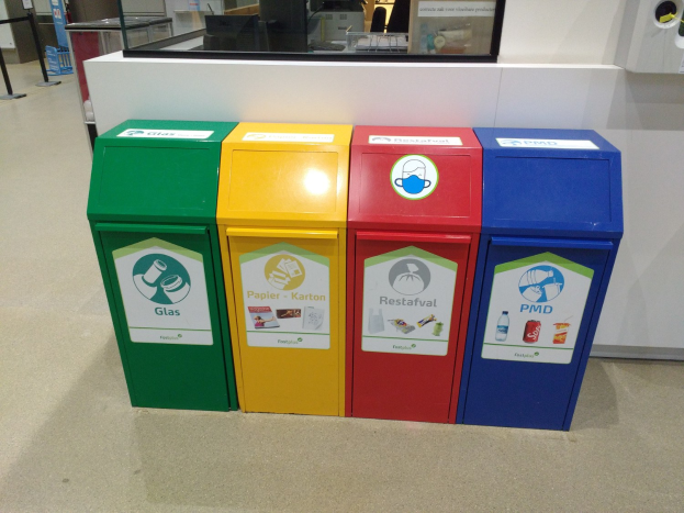 Vier bunte Mülltonnen in einer Reihe auf dem Boden, mit einer Theke, einem Monitor und anderen Gegenständen im Hintergrund und einer Person links daneben.